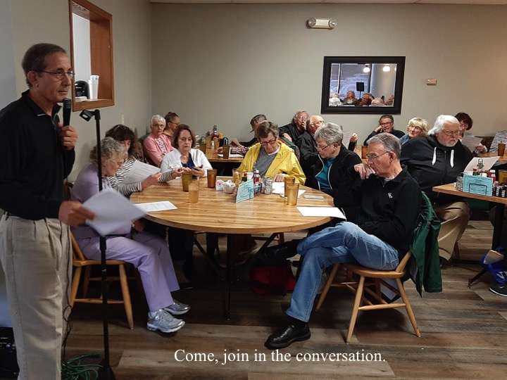 A man presents to a room full of people seated around tables