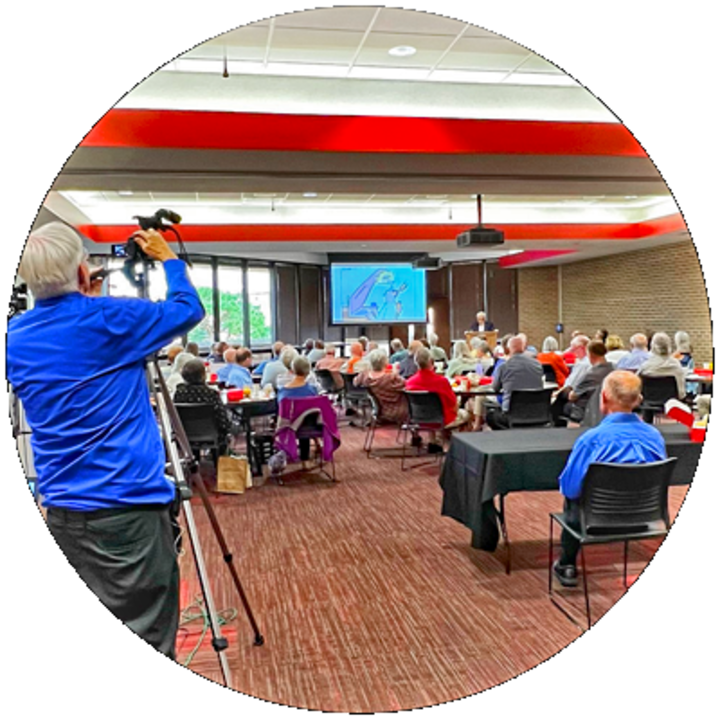 a man records a room full of people seated around tables